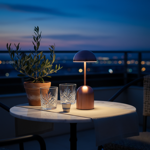 Table with a lamp, glasses, and a plant on a balcony at night.