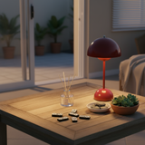 Red lamp on a wooden table with dominoes, a plant, and a bowl of snacks in a room with plants outside.