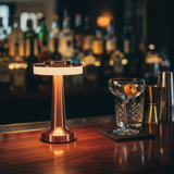 Cocktail glass with a slice of orange on a bar counter next to a lamp.