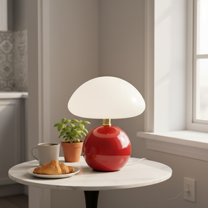 Red table lamp with a white shade on a small round table next to a cup, croissant, and plant.